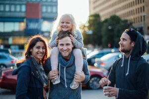 Image of a smiling family standing on a street in Helsinki. The man is carrying a little blond girl on his shoulder and next to the family stands another man with a coffee in his hand